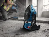 A person wearing safety equipment operates a power drill beside a portable jobsite fan.