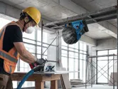 A person wearing safety equipment uses a power saw on wood near a portable fan.