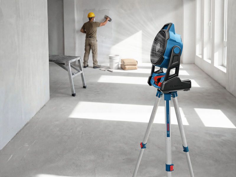 A person wearing safety equipment smooths a wall while a portable fan stands on a tripod.