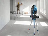 A person wearing safety equipment smooths a wall while a portable fan stands on a tripod.