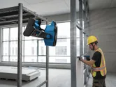 A person wearing safety equipment operates a power tool near a mounted portable fan.
