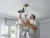 A person wearing safety equipment installs a chandelier using a cordless drill on a ladder.