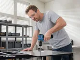 A person wearing safety equipment drills into a tabletop using a cordless drill.