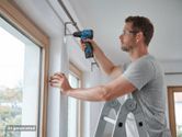 A person wearing safety equipment uses a cordless drill to install a curtain rod.