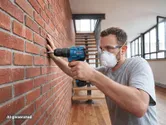 A person wearing safety equipment drills into a brick wall with a cordless impact drill.