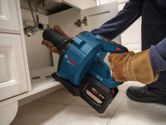 A person wearing safety equipment uses a cordless drain cleaner under a sink.