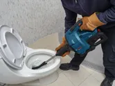 Person wearing safety equipment uses a power tool to unclog a toilet.