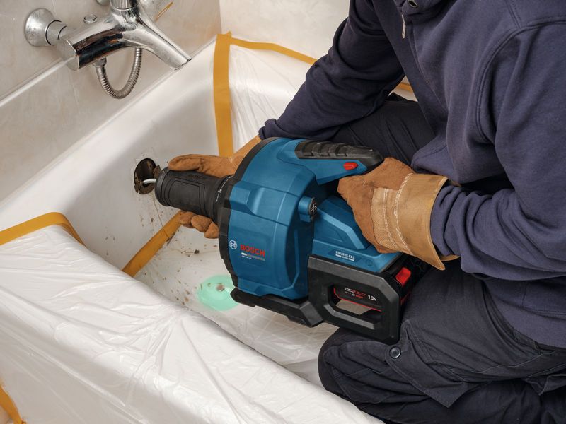 A person wearing safety equipment uses a drain cleaning tool in a bathtub.