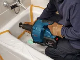 A person wearing safety equipment uses a drain cleaning tool in a bathtub.