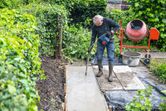 A person wearing safety equipment uses a power tool to compact concrete on a garden path.