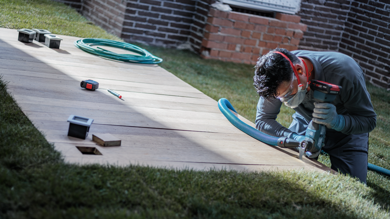 A person wearing safety equipment drills into a wooden panel on grass.