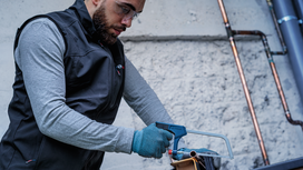Person wearing safety equipment cuts a metal pipe with a hacksaw.