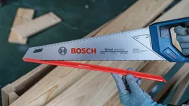 Person wearing safety equipment holds a hand saw above stacked wooden planks.