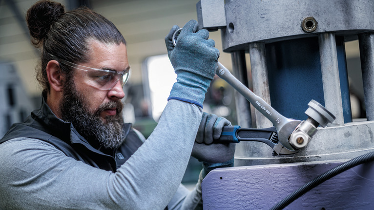 Person wearing safety equipment tightens a large bolt with a wrench on industrial equipment.