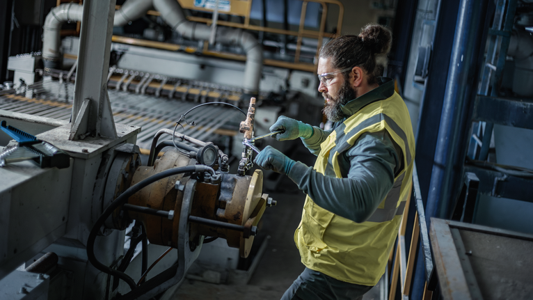 Person wearing safety equipment uses a power tool to adjust machinery in a factory.