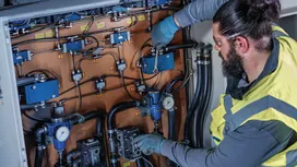 A person wearing safety equipment adjusts hydraulic controls inside an industrial panel.