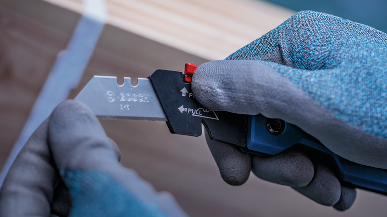 Person wearing safety equipment adjusts a utility knife near a wooden surface.