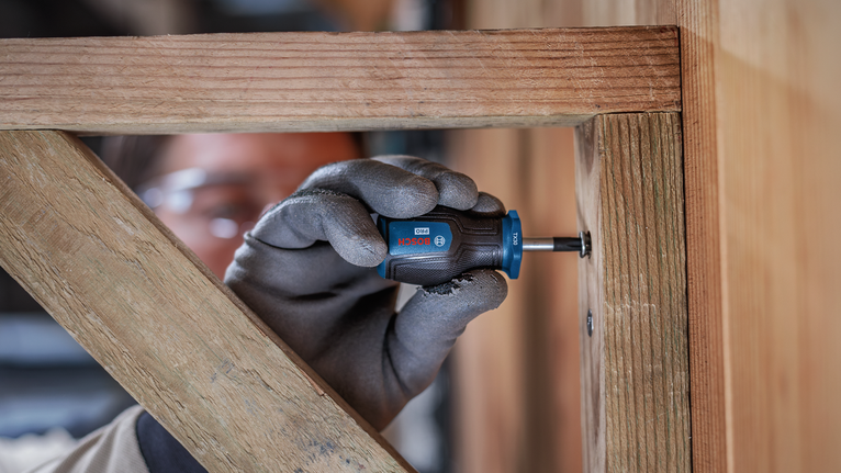 Person wearing safety equipment uses a screwdriver to fasten a screw in a wooden frame.
