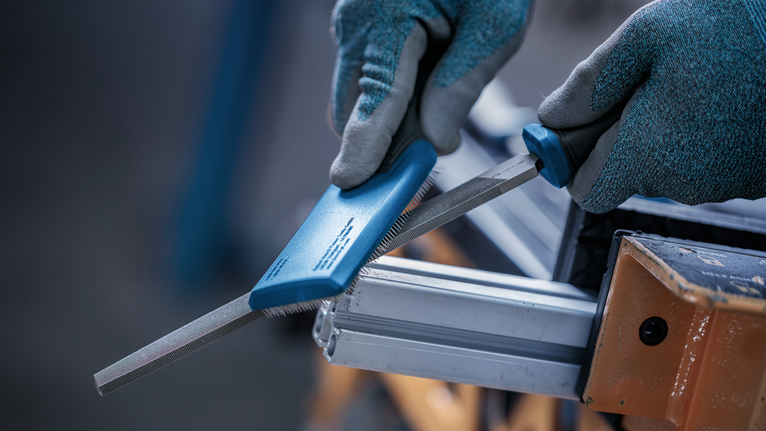 Person wearing safety equipment uses a file to smooth a metal bar.