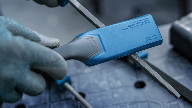 Person wearing safety equipment files a metal rod with a blue-handled tool.
