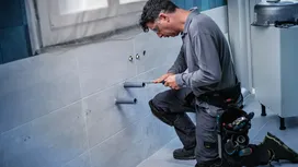 Person wearing safety equipment working on wall pipes in a tiled bathroom.