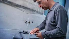 Person wearing safety equipment files a pipe protruding from a tiled wall.