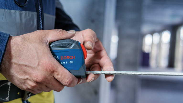 Person wearing safety equipment measuring with a retractable tape in a building.