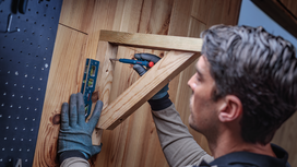 Person wearing safety equipment uses a screwdriver to attach a wooden frame to a wall.