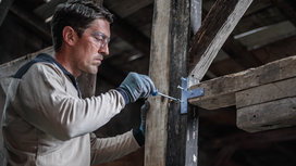 Person wearing safety equipment drives a screw into a wooden beam bracket.