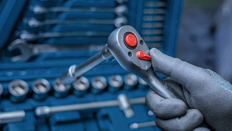Person wearing safety equipment holds a ratchet handle in front of a tool set.