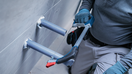 Person wearing safety equipment cuts a wall-mounted pipe with a hacksaw.