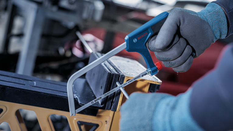 Person wearing safety equipment cuts a metal bar with a bow saw.
