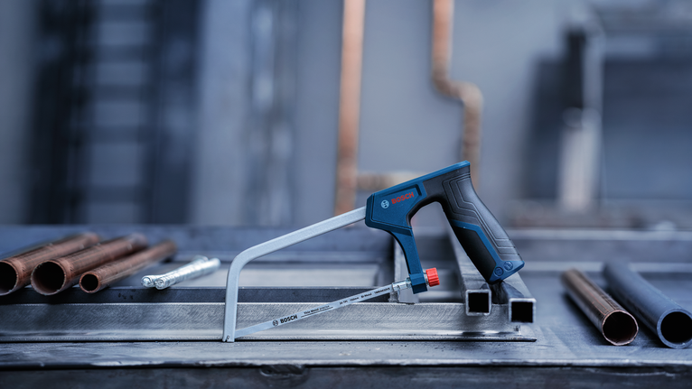 Hacksaw with a black and blue handle placed on a metal work surface next to pipes.