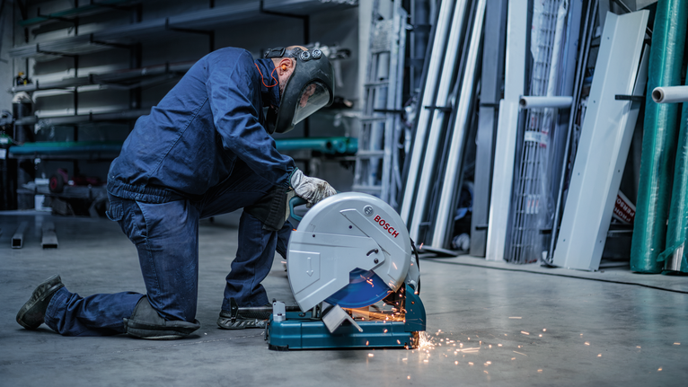 A person wearing safety equipment uses a metal chop saw, producing sparks.