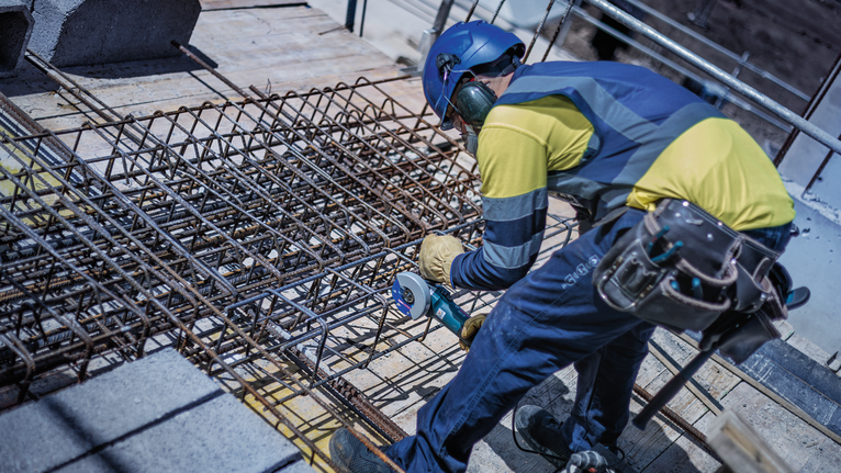 Person wearing safety equipment uses a power tool to cut rebar on a construction site.