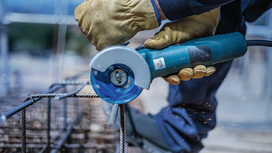 Person wearing safety equipment cuts metal rebar using a blue handheld angle grinder.