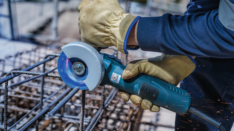 Person wearing safety equipment uses angle grinder to cut metal rebar.