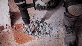 Person wearing safety equipment breaking concrete with a demolition tool.
