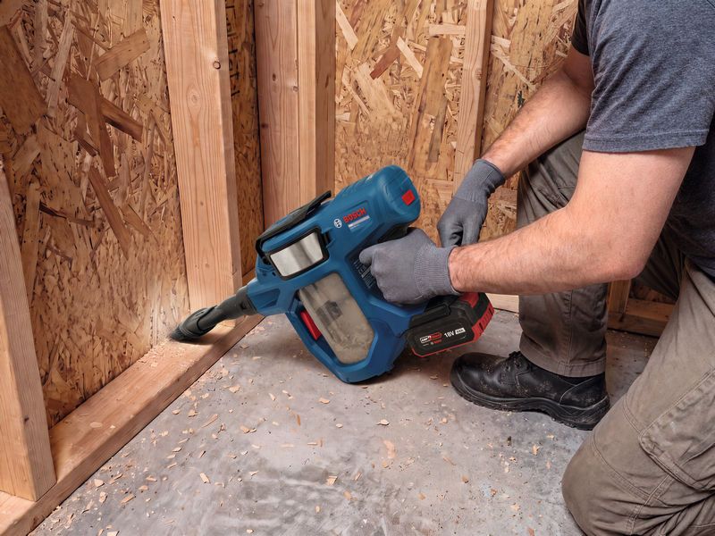 A person wearing safety equipment uses a cordless vacuum cleaner to clean wood debris.
