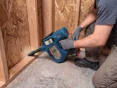 A person wearing safety equipment uses a cordless vacuum cleaner to clean wood debris.
