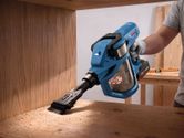 A person wearing safety equipment uses a cordless dry vacuum cleaner to collect wood dust.