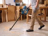 A person wearing safety equipment uses a cordless vacuum cleaner to clean wood shavings on a workshop floor.