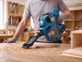 A person wearing safety equipment uses a cordless vacuum cleaner to remove wood dust.