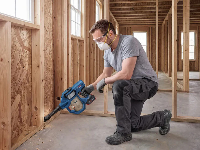 A person wearing safety equipment uses a cordless dry vacuum cleaner on a construction site.