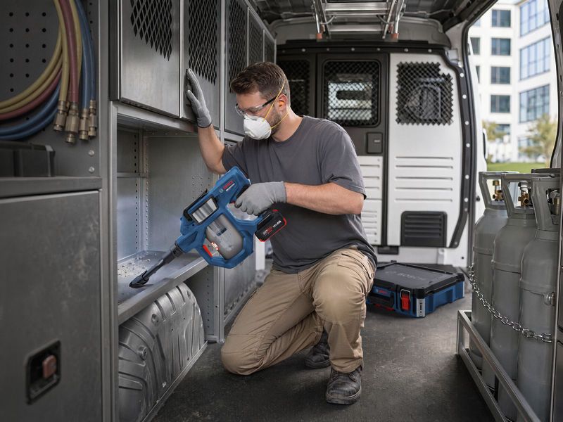 A person wearing safety equipment uses a cordless dry vacuum cleaner inside a work van.