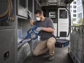 A person wearing safety equipment uses a cordless dry vacuum cleaner inside a work van.