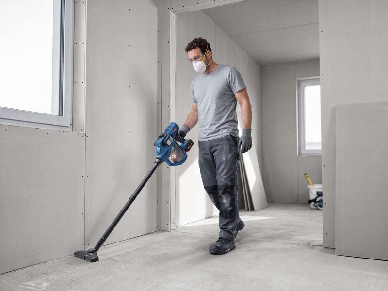A person wearing safety equipment cleans a construction site floor with a cordless dry vacuum cleaner.