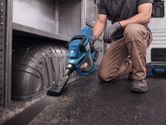 A person wearing safety equipment uses a cordless dry vacuum cleaner on a vehicle floor.