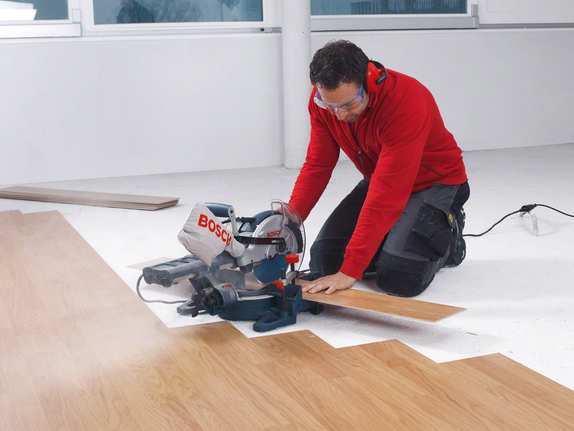 A worker wearing safety equipment cuts laminate flooring with a sliding miter saw.