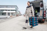 A person wearing safety equipment carries stacked toolboxes beside a work van at a construction site.